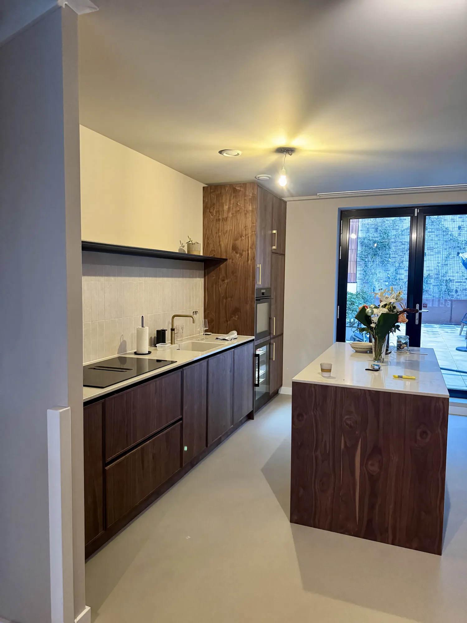 Keuken in noten met een kookeiland met een licht stenen blad en een hoge kastenwand met oven