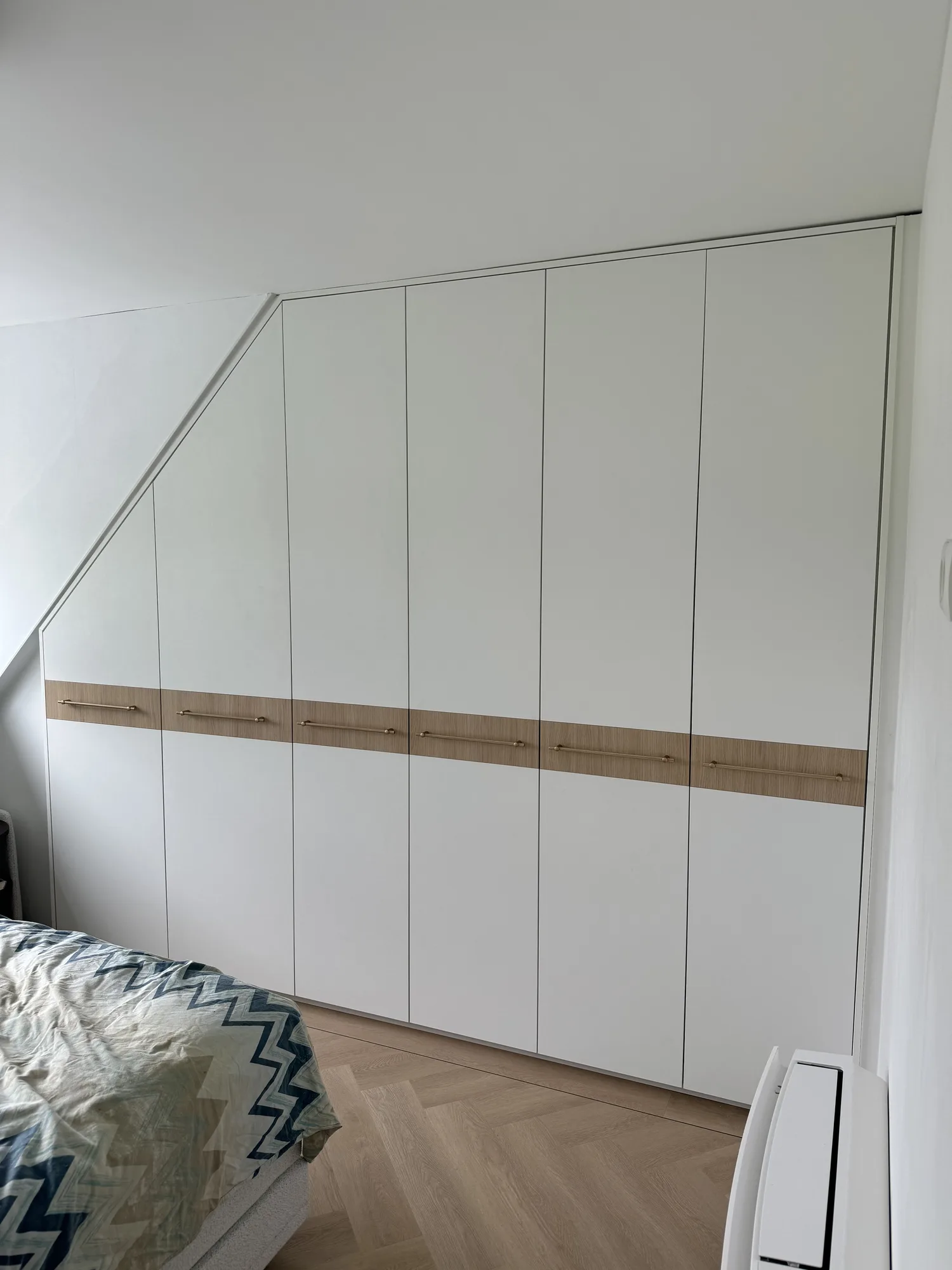 Witte kledingkast onder een schuin dak, met een strook in eiken als greep